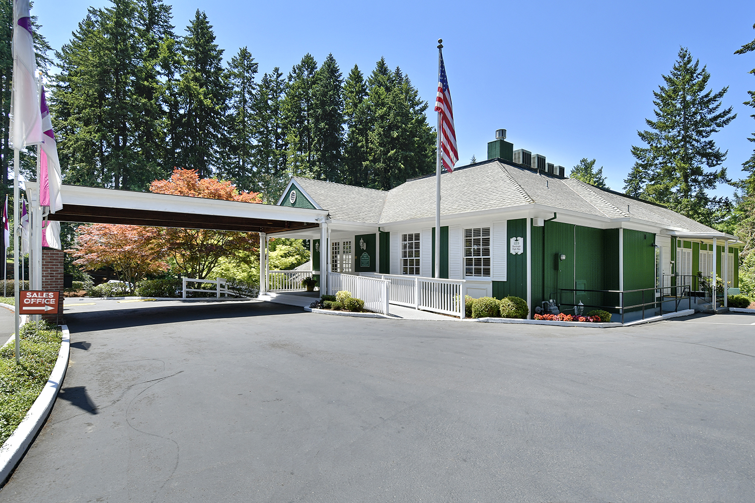 Heritage Village, an all age manufactured home community in Beaverton. Sales office inside green building and marked with red Sales Office sign pointing to the right. Large American flag on tall flagpole. Very tall trees in background. Clean, paved driveway up to building.