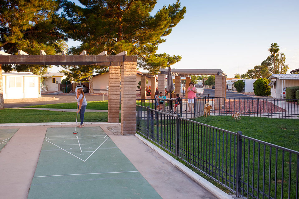 Woman plays shuffleboard next to fenced in dog park while dogs run free and a family watches on from the picnic benches