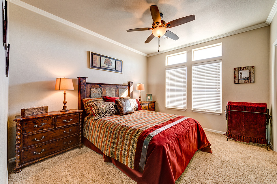 Red, tan, and brown theme bedroom. Dark wood bed frame and night stands with lamps. Ceiling fan combined with light and large window on the wall for natural light.