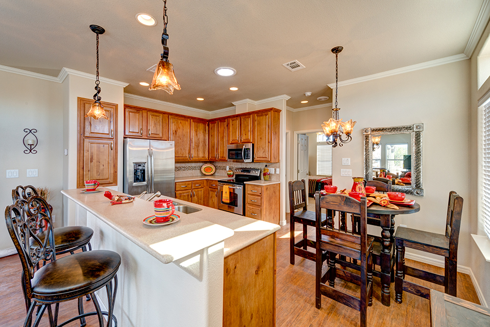 Updated L shaped kitchen with wood cabinets, stainless steel appliances, and white counter tops. Hanging ceiling lights over seating areas. Ample eating space on either the elevated counter or on the circular kitchen table, all equipped with tall dark colored bar chairs.