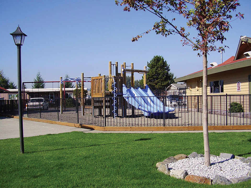 Outdoor playground enclosed by gate. Sand covers the ground throughout and a grass area outside of the gates.