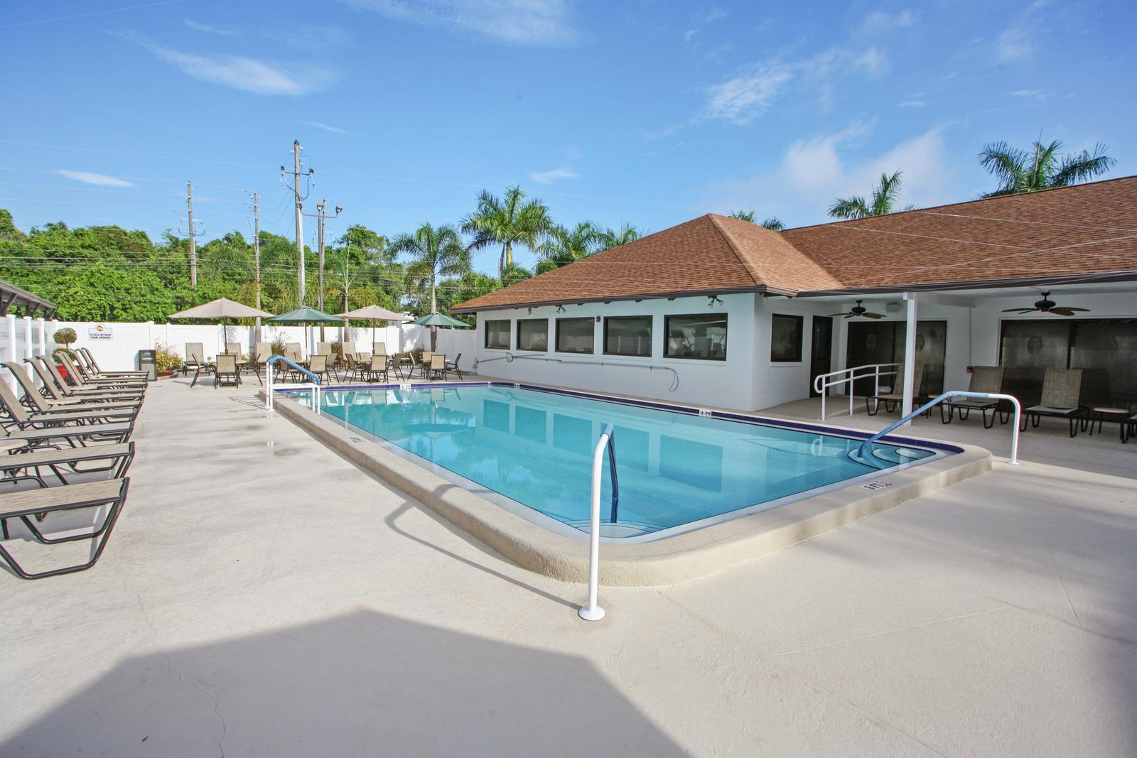Calm swimming pool with lounge chairs and patio furniture with chairs, tables, and umbrellas. located outside clubhouse.