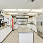 Beautiful modern commercial kitchen in clubhouse with all stainless steel appliances and plenty of white cabinets for storage. Long island with sink in the middle.