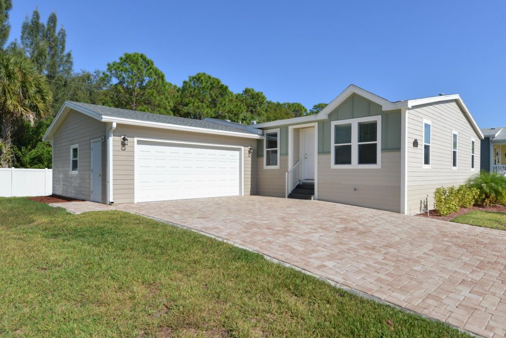 New homes are for sale at Lamplighter Village, an active 55 plus manufactured home community. Front of home with attached 2 car garage. Long driveways of pavers. Green grass and small plants for front landscape. Lush green trees in the background