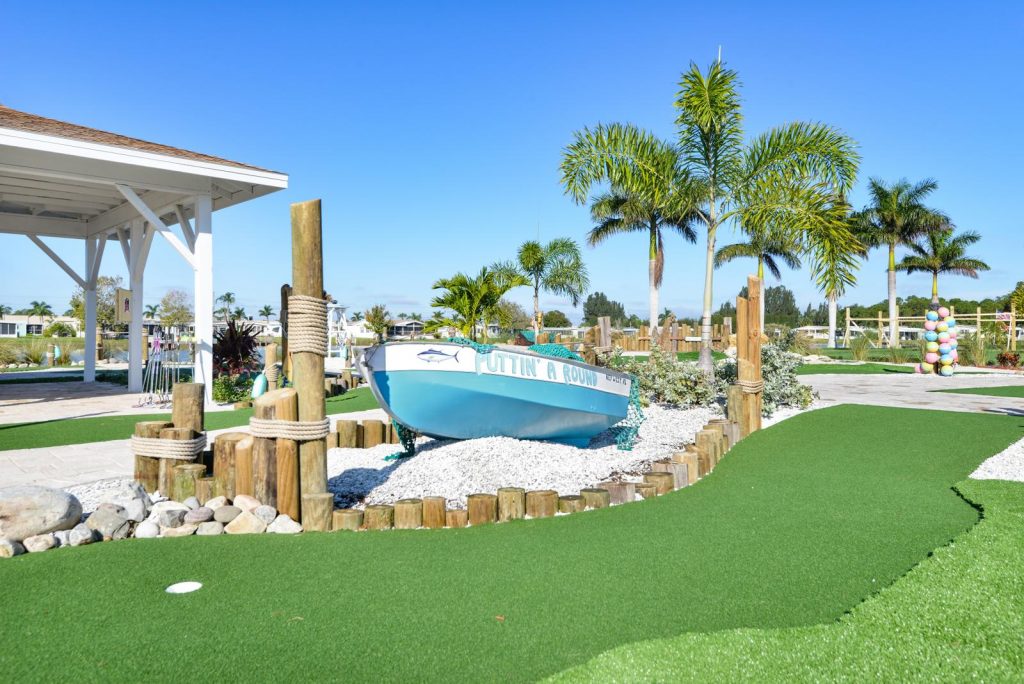 Mini golf course with clean greens. A blue fishing boat sits upon white rocks with palm trees along the course.