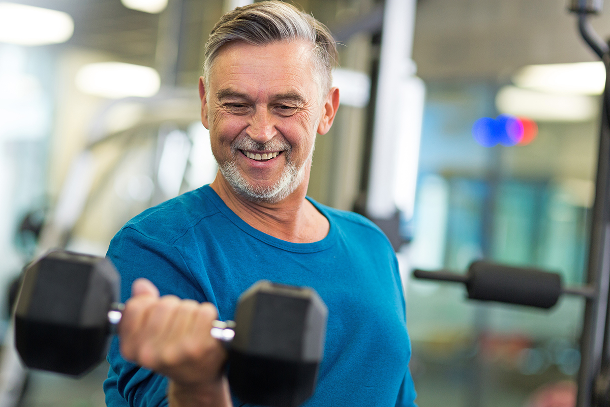 A 55+ man works out in fitness center and curls arm with free weight.