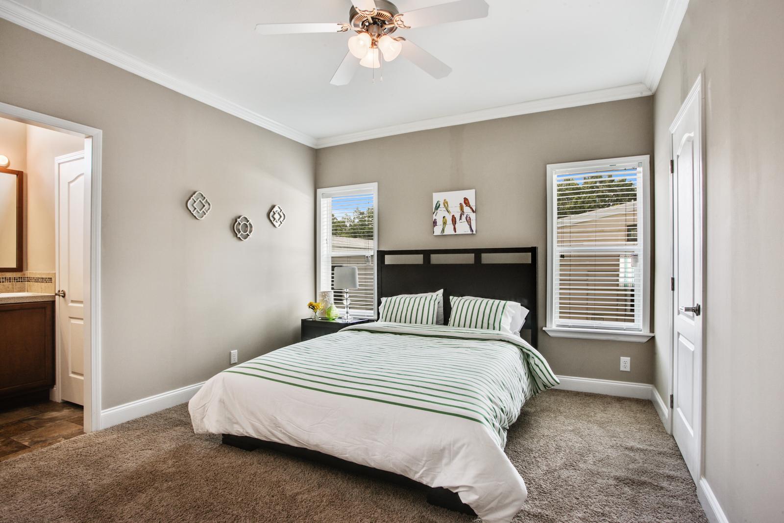 Bedroom with attached bathroom. Light gray walls with crown molding on top and bottom. Two windows. Queen sized bed with dark wood headboard and end table. Light brown carpeting.