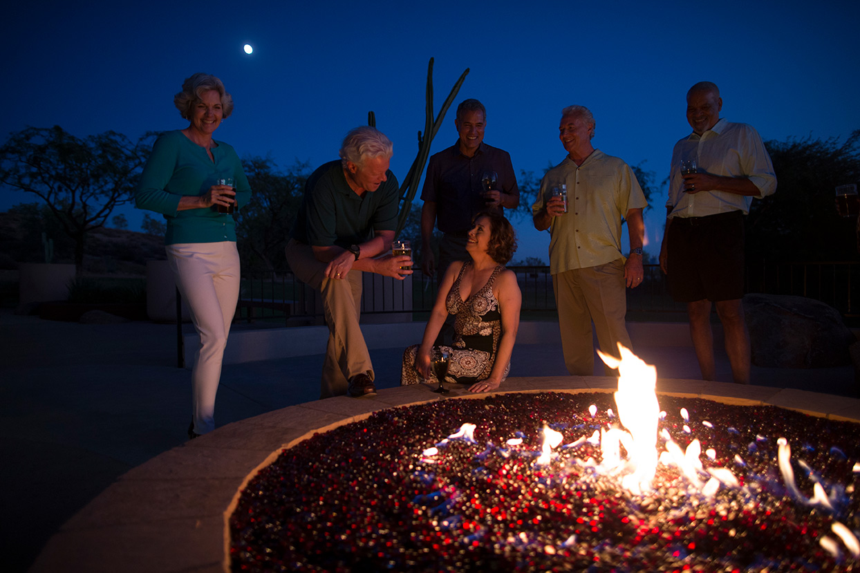 At night around the large lit fire pit, four men and two women enjoy a drink and chat while under the moon light