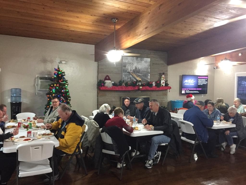 Community holiday party hosted in the community hall. 55 plus aged residents sit and enjoy dinner meals while mingling with one another. Community hall is decorated for the holidays with a Christmas tree and red and green decor over the fireplace ledge.