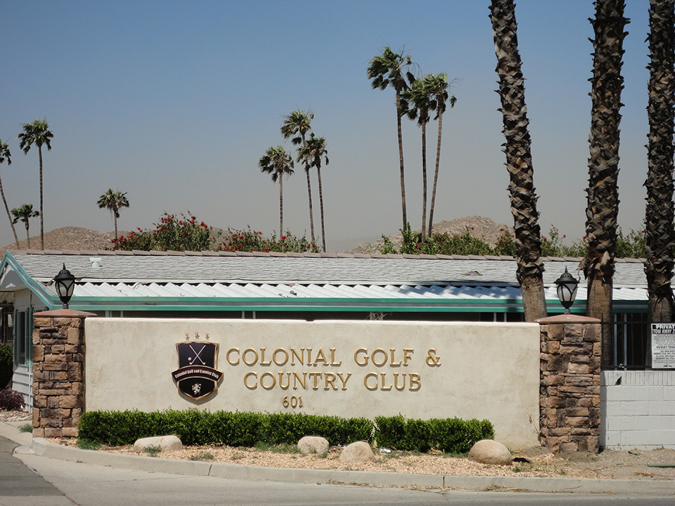 Entrance to the beautiful 55+community, golf course and country club. Greeted by a beige, cement wall labeled with Colonial Golf and Country Club, with light brick pillars on each side and palm trees in the background.