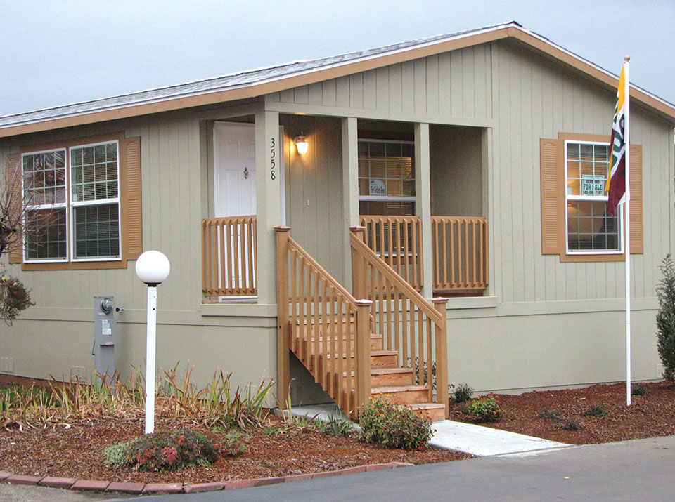Manufactured home painted sage green outside with tan trim and window shutters. Covered front porch. Maintained landscape with a few flowers around the white lamppost.