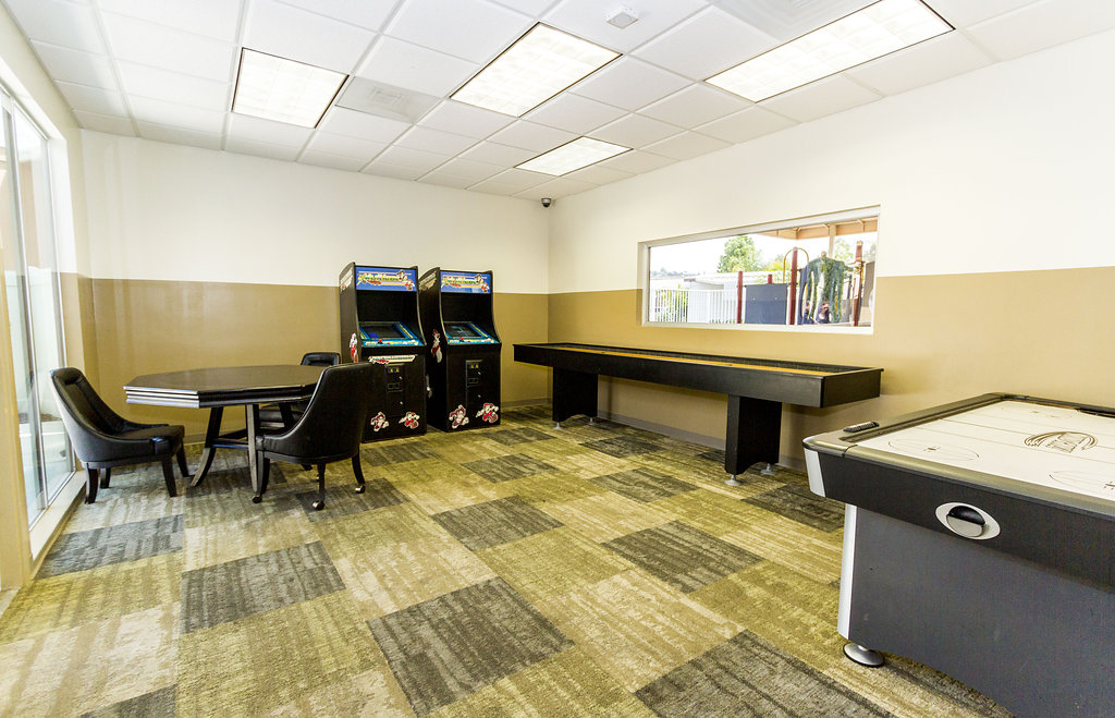 Game room including an ice hockey table, a poker table with chairs, shuffleboard table, and two arcade game systems. Small open space for residents to enjoy games with friends and families.