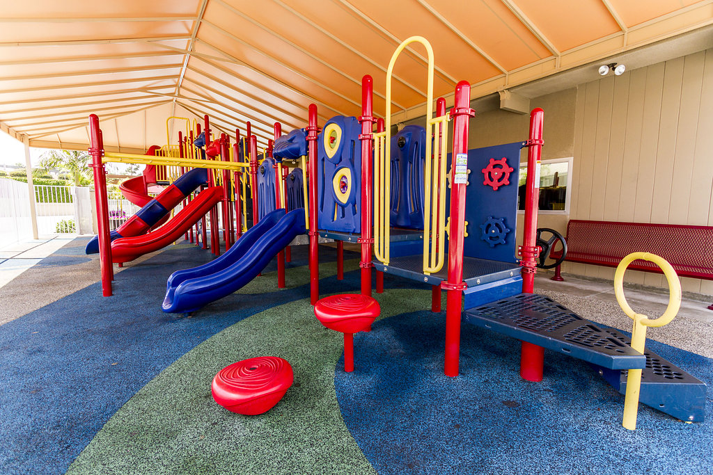 Vibrant red, blue, and yellow outdoor community playground. Covered by large steel metal carport/shelter/ shade with ample, open space for play. Rubber playground flooring throughout to ensure safety.