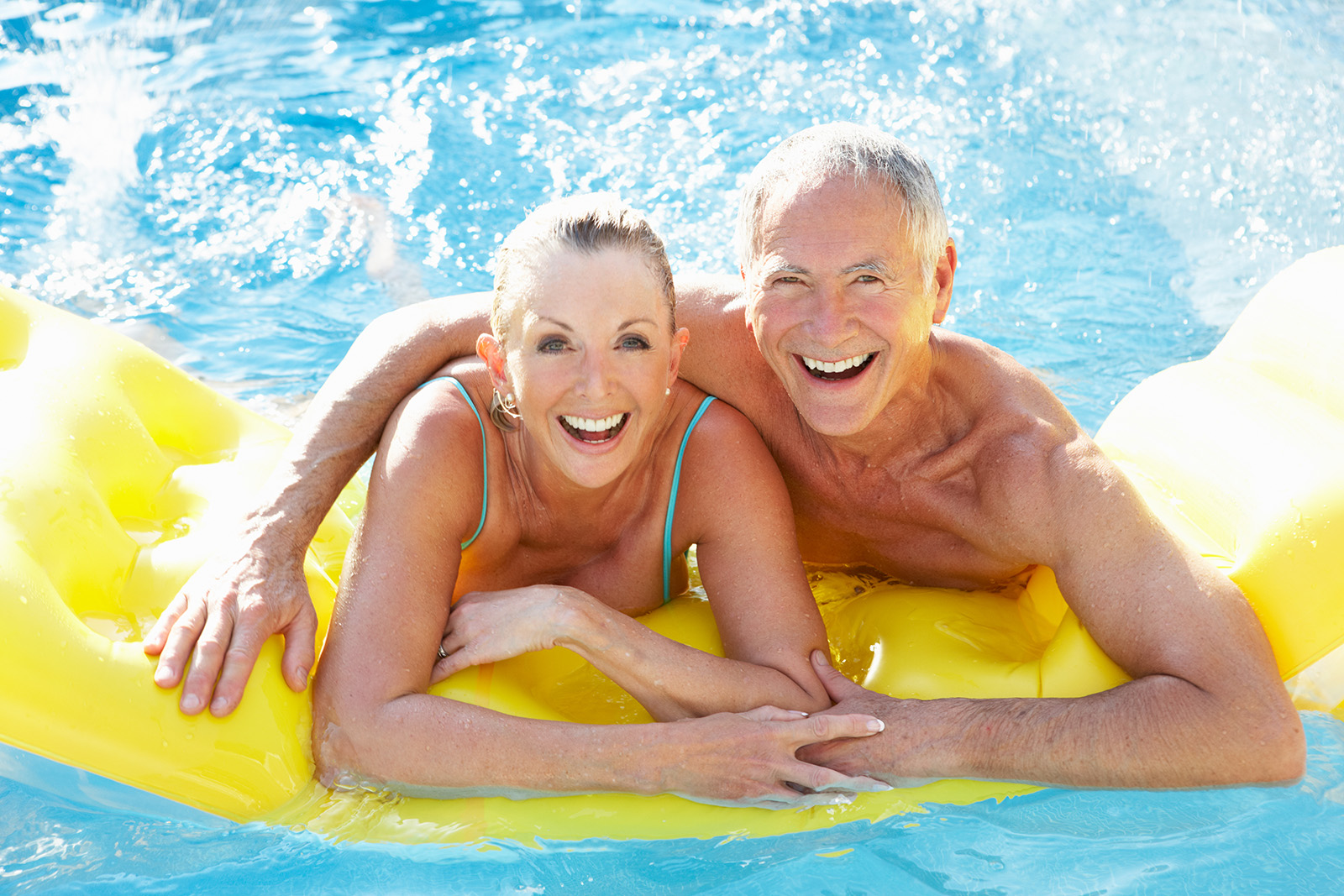 A mature, older couple enjoy their raft in the pool. The man and woman hold hands and smile.