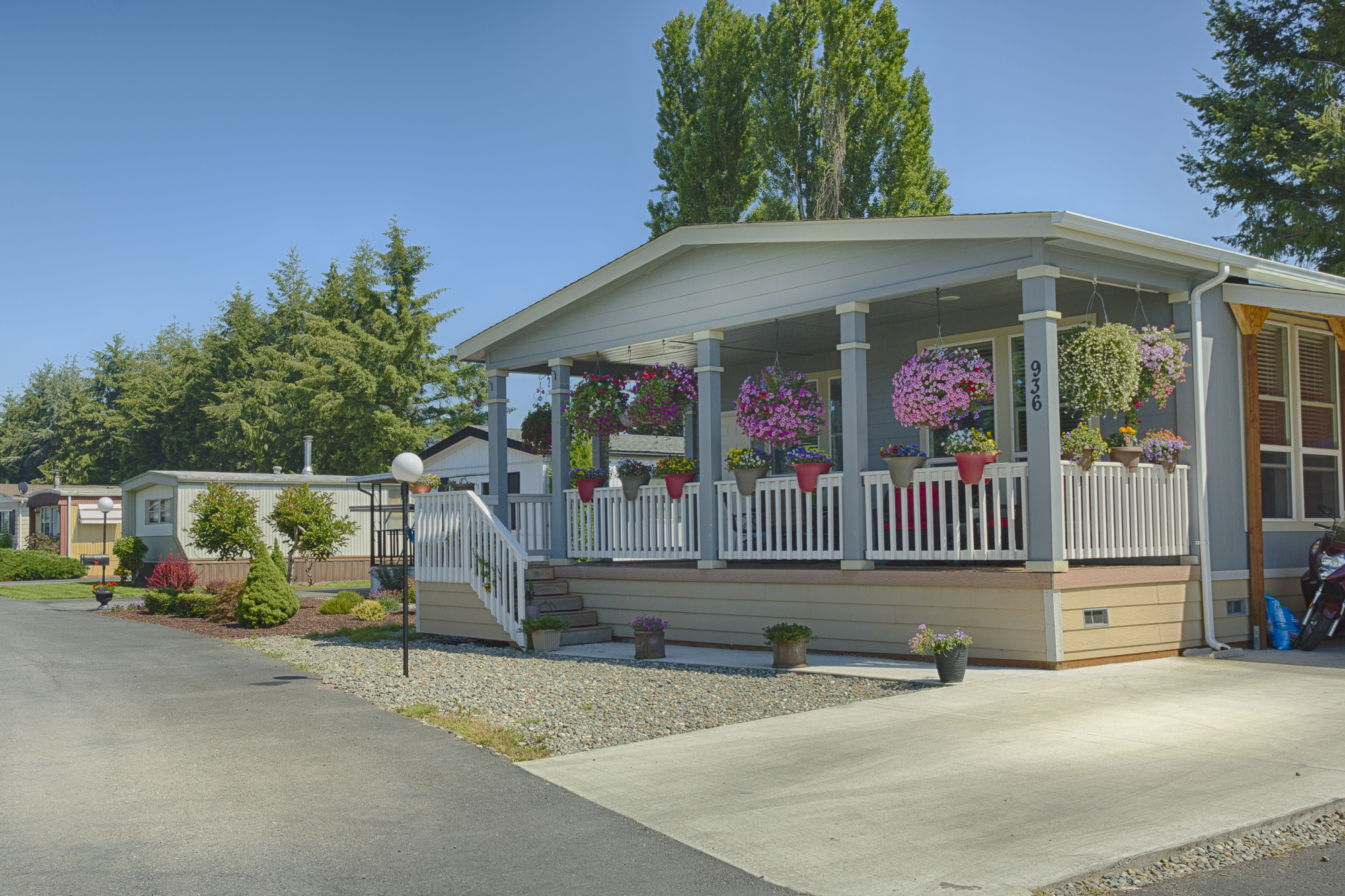 Beautiful manufactured home with large covered front porch. Colorful potted flowers hang around the perimeter of the porch.