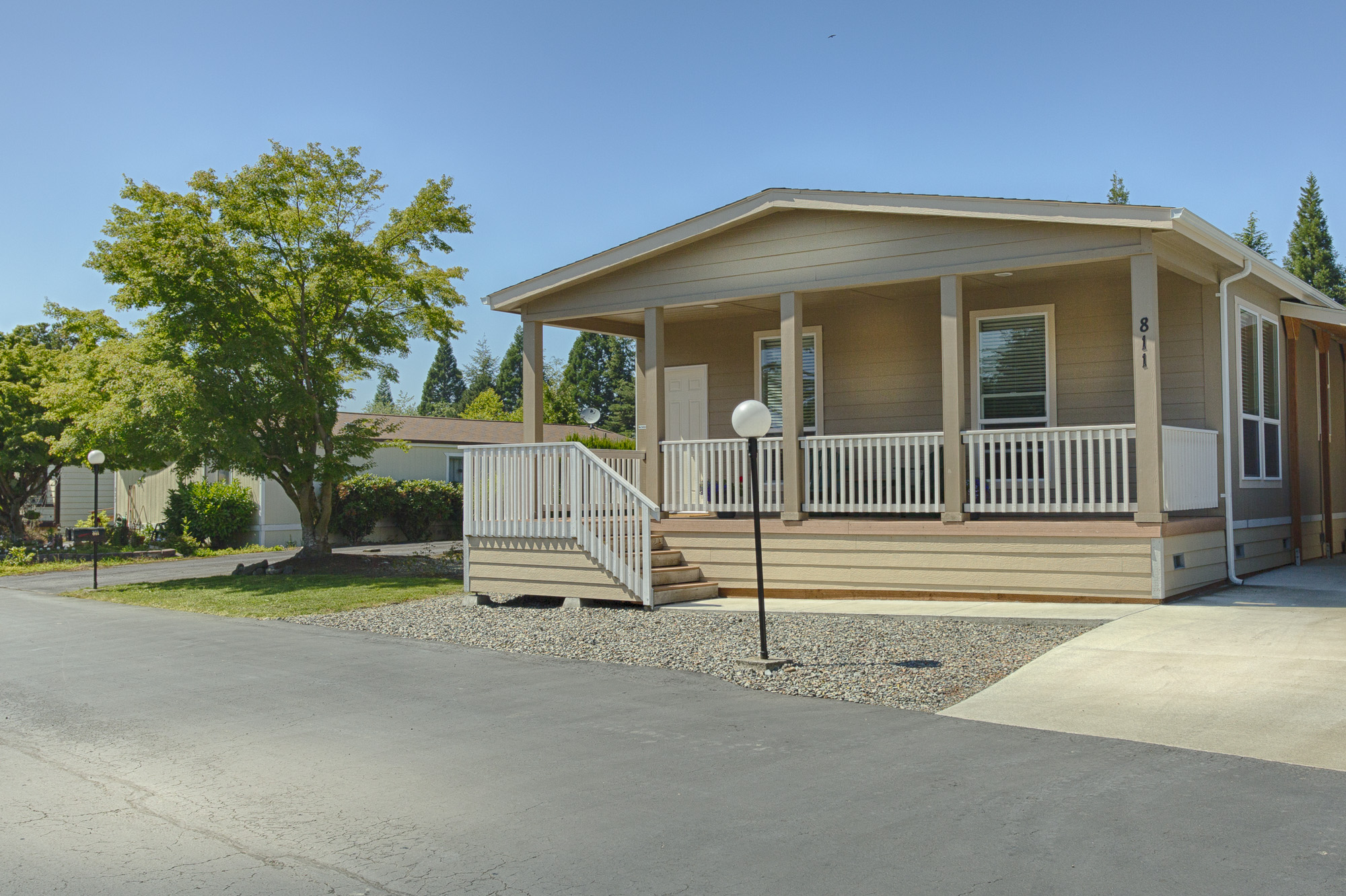 Beige colored manufactured home with small staircase leading up to long covered front porch. Very well-maintained front landscape for a clean look.